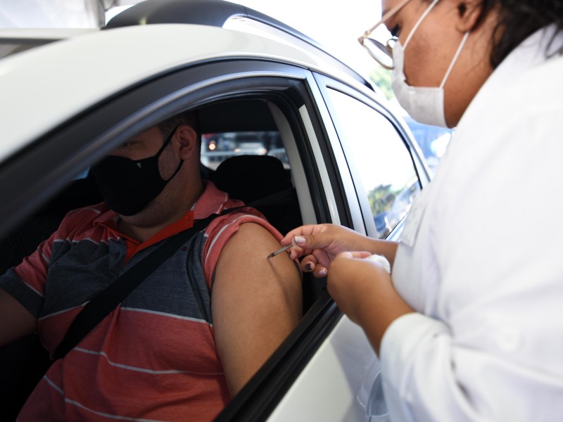 Foto de vacinação contra a Covid em posto drive-thru. Uma profissional da saúde está aplicado a vacina no braço de um homem, que está dentro do carro, sentado no banco do motorista